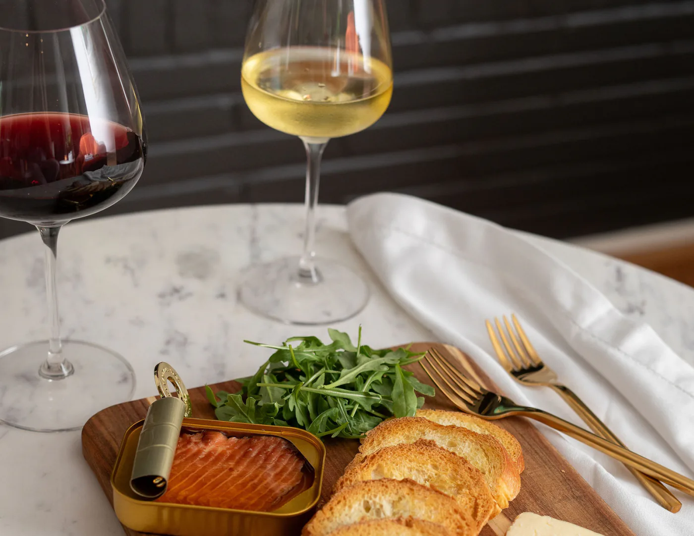 A glass of red and a glass of white wine on a marble table behind a wooden board of smoked trout, arugula, sliced baguette and gold cutlery.
