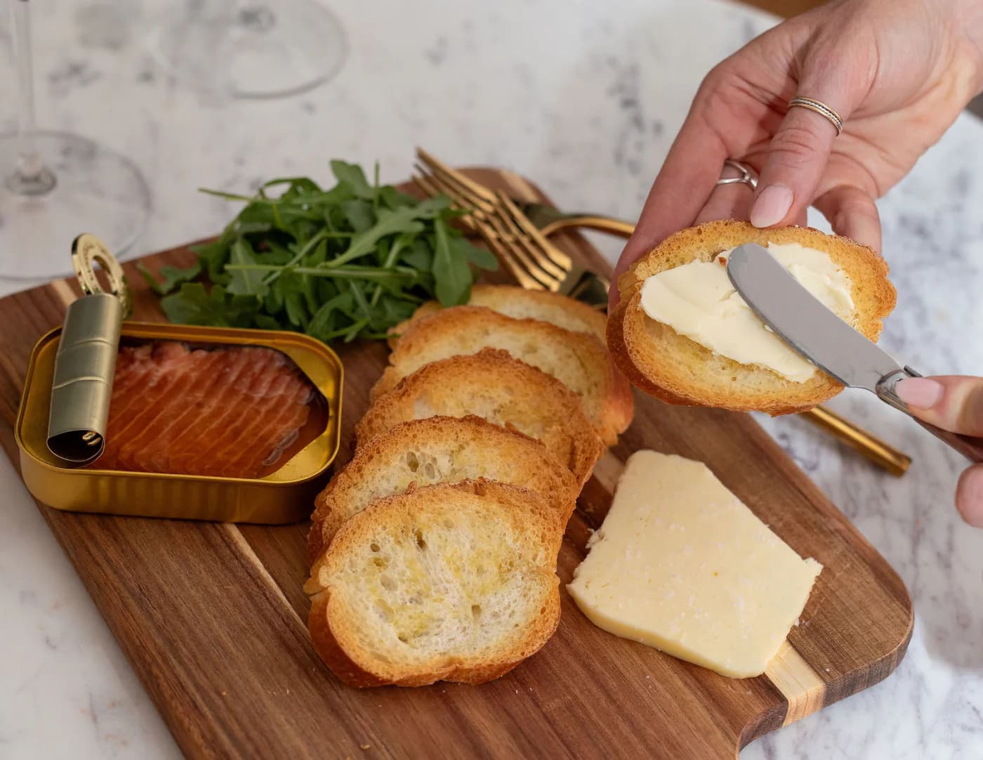 A wooden board on a marble table holding a tin of smoked trout, arugula, sliced baguette and a wedge of hard cheese, with a hand buttering a slice of toast.